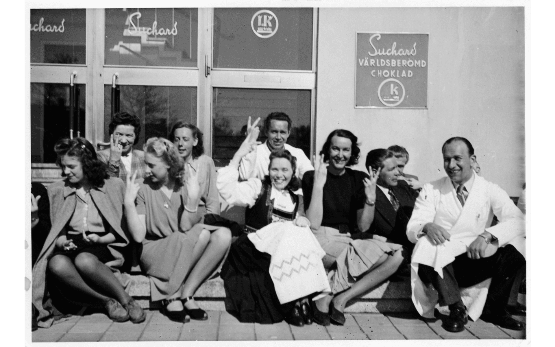 Delar av personalen på Suchards Chokladfabrik utanför entrén till fabriken som var belägen i kvarteret Konfektasken. Bilden är troligen tagen i samband med fredsdagen 7 maj 1945 då alla gör fredstecken. Fotografering - maj, 1945