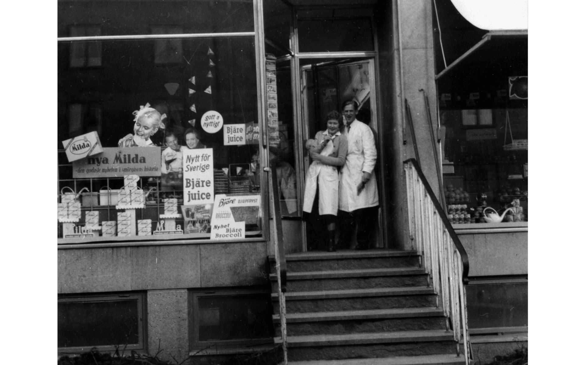 Bild på en man och en kvinna som står vid ingången till Konsumbutik på Sveagatan 12. Fotografering - 1950-1960.