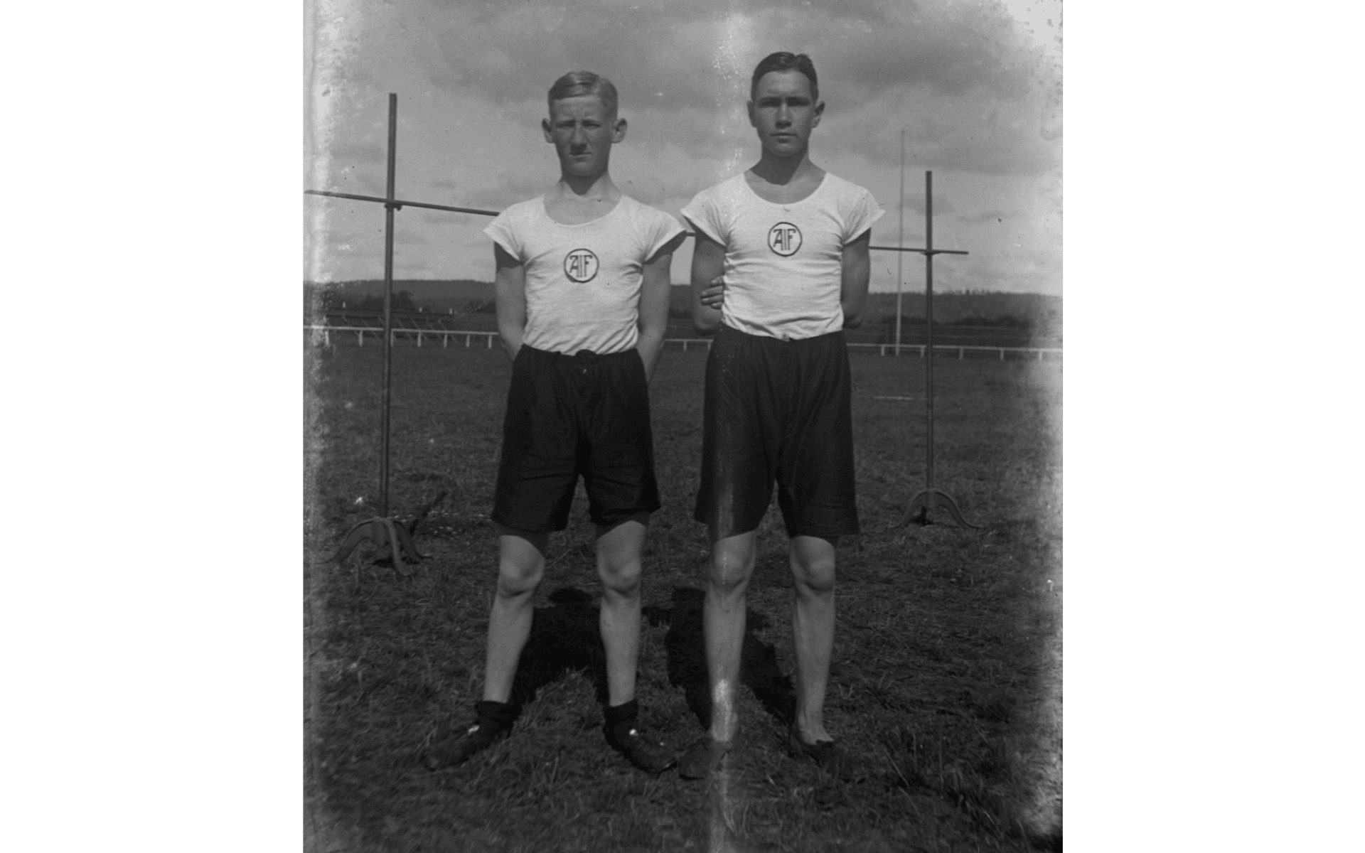 Gösta Larsson och Halvar Olsson fotograferade framför ett höjdhoppshinder på idrottsplatsen. Båda bär mörka shorts och vita t-shirts med AIFs (Alingsås Idrottsförenings) märke. Fotografering - 1916.
