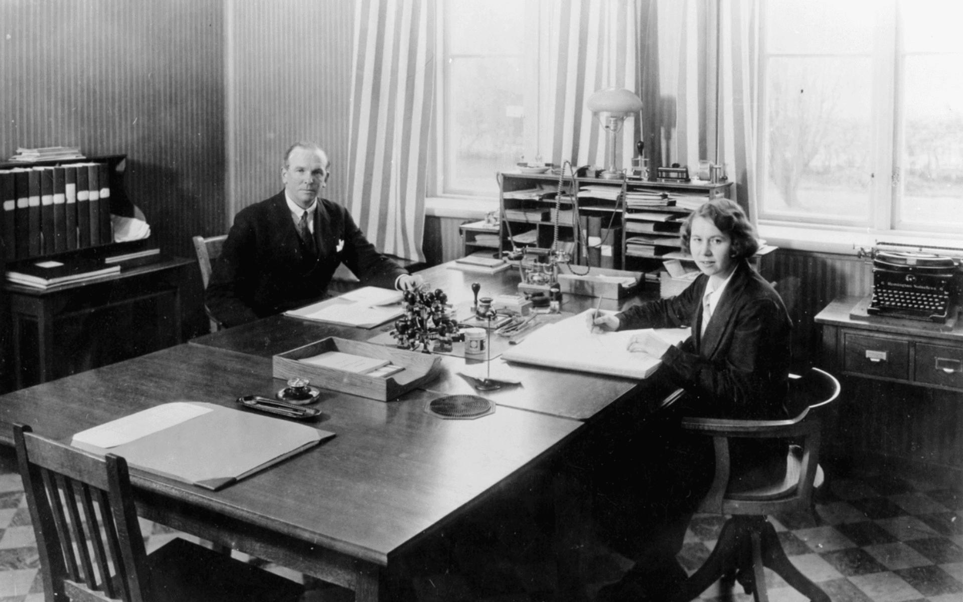 Kontorsrum med en man och en kvinna på var sin sida av ett större skrivbord. Inifrån Alingsås lasarett. Från 1940-talet.